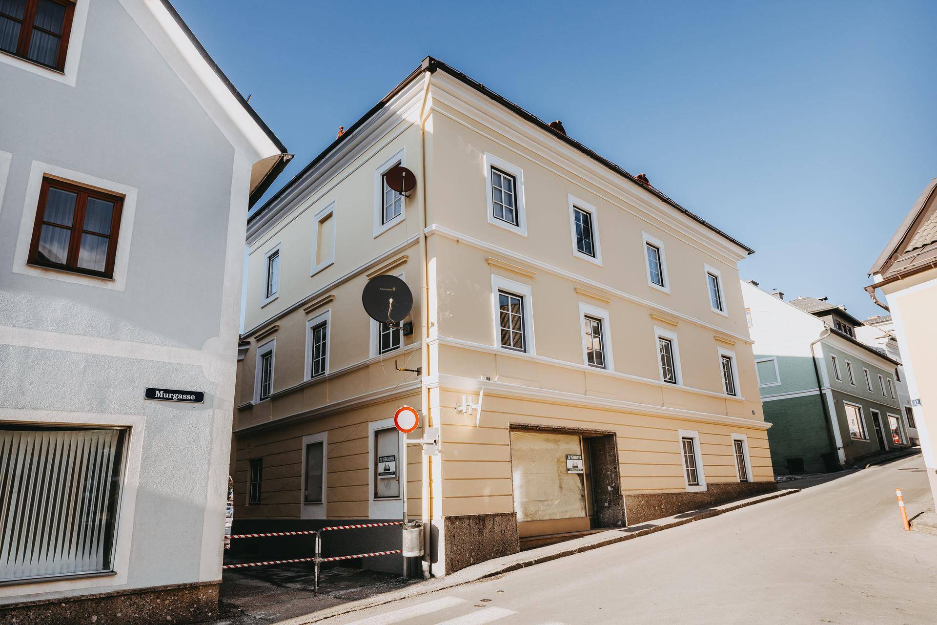 Eckgebäude mit gelber Fassade und traditionellen Fenstern an einer leicht ansteigenden Straße.