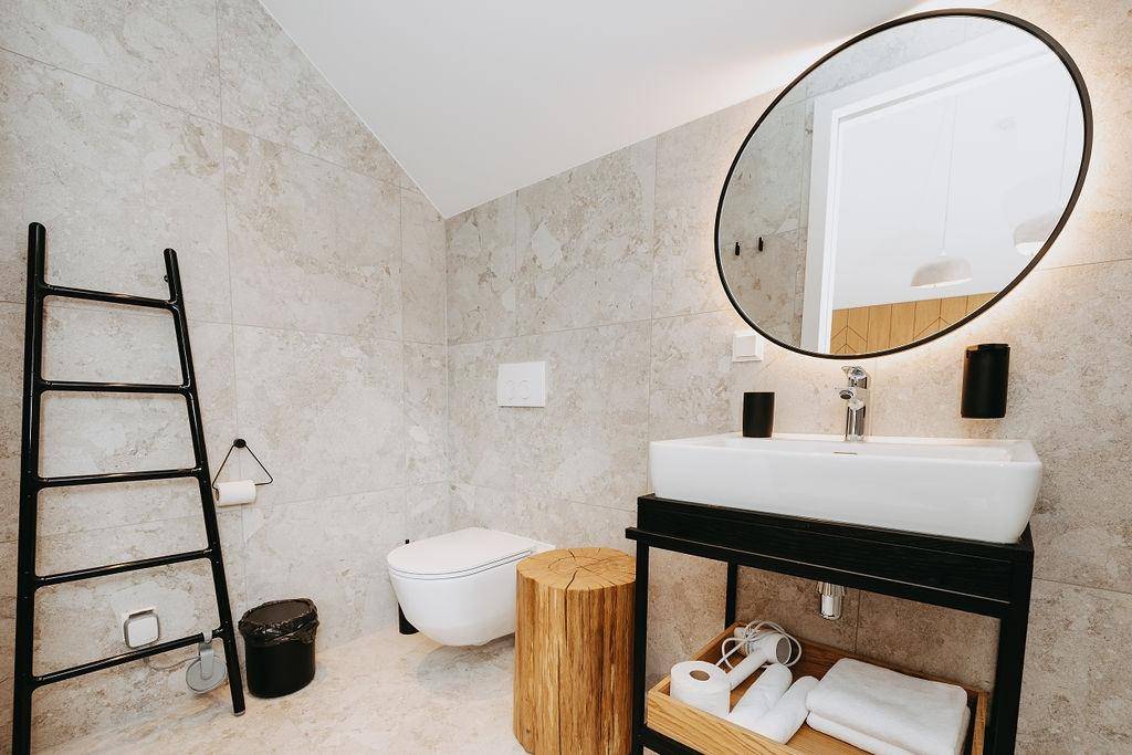 Modernes Badezimmer mit hellen Fliesen, wandhängendem WC und ovalem Spiegel mit Beleuchtung.