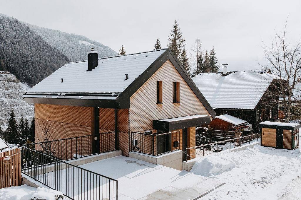 Vorderansicht des modernen Holzhauses im Winter mit schneebedeckter Auffahrt und alpiner Kulisse.