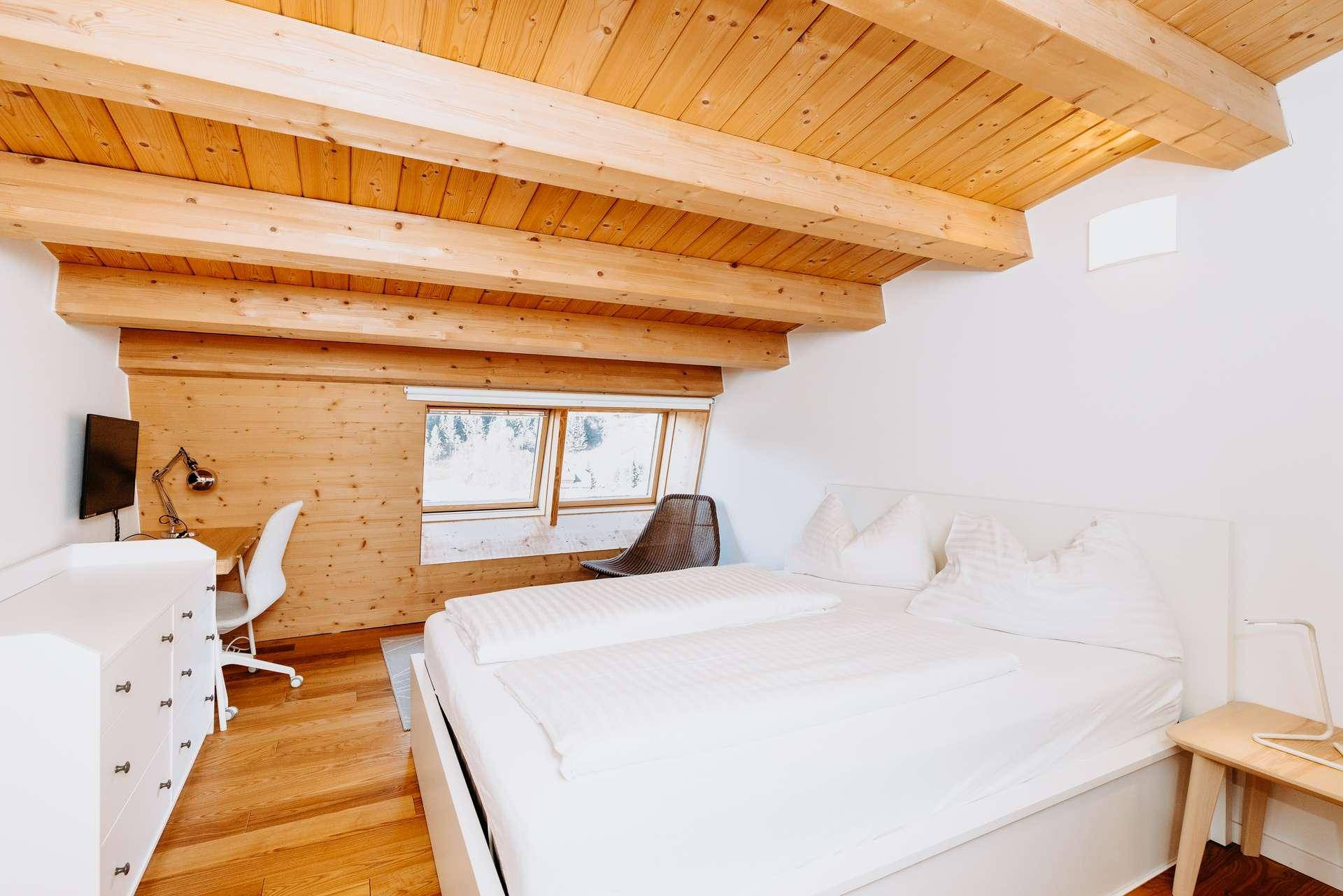 Gemütliches Schlafzimmer mit Holzbalken, Holzwänden und Arbeitsbereich am Fenster.