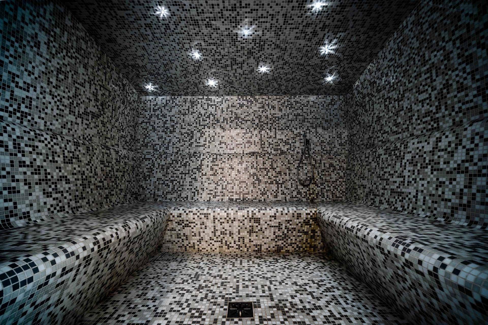 Dampfbad oder Hamam mit Mosaikfliesen an Wänden und Bänken, beleuchtet von Sternenlichtern an der Decke.