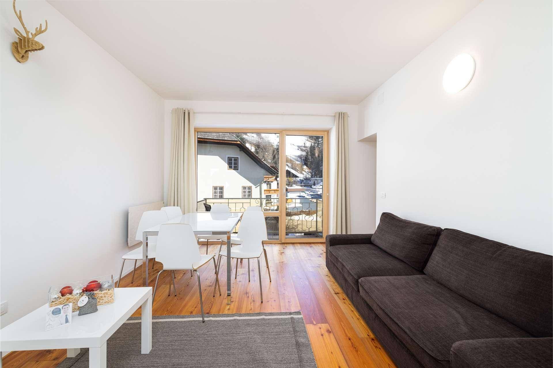 Heller Wohn-Essbereich mit Holzboden, dunklem Sofa, Esstisch und Zugang zu einem Balkon.