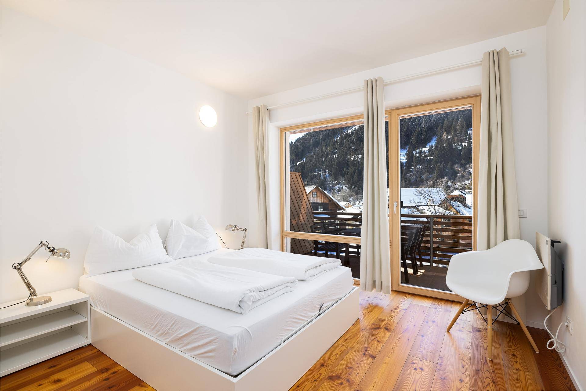 Schlafzimmer mit weißem Bett, Holzboden und großen Fenstern mit Blick auf eine verschneite Berglandschaft.