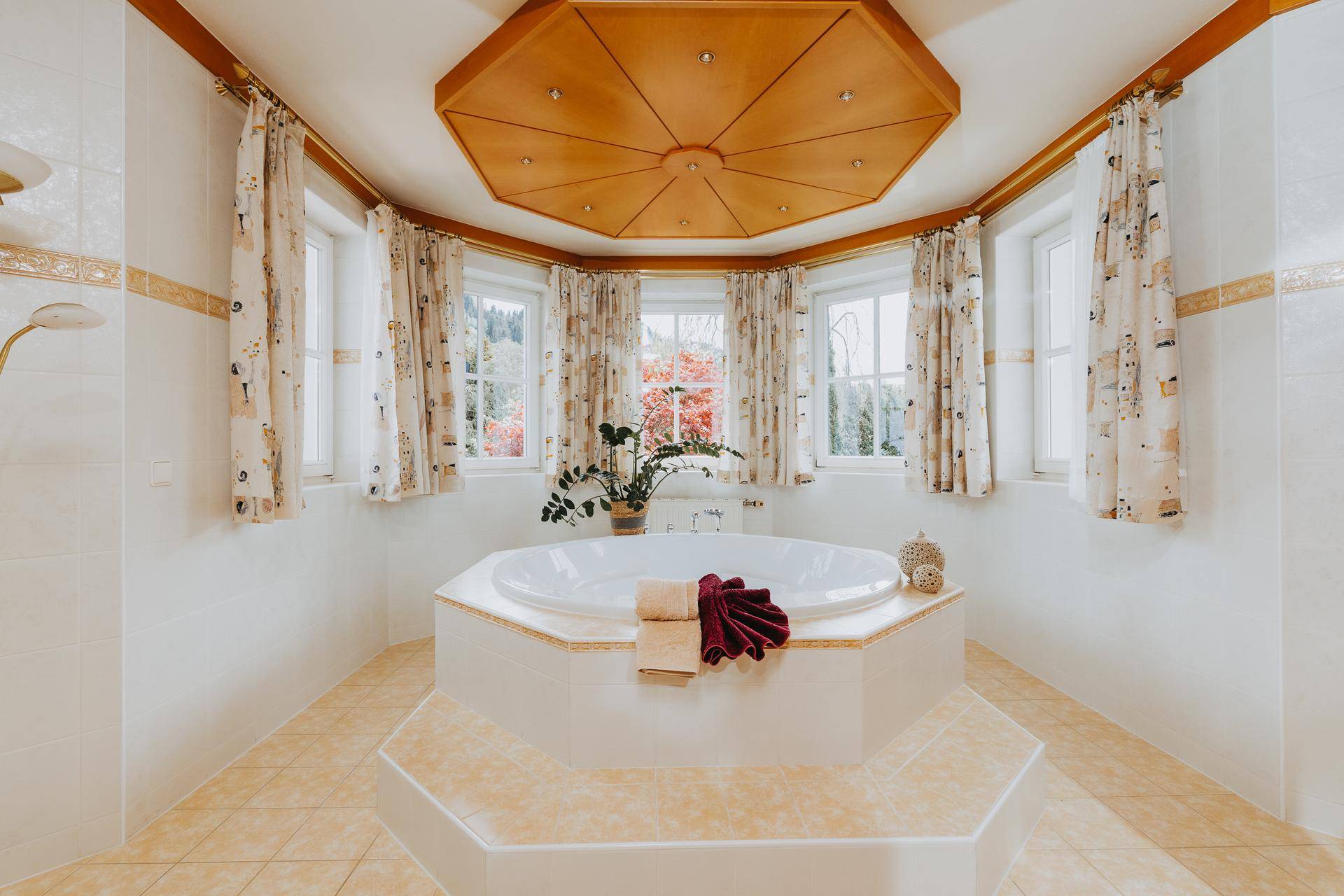 Großes Badezimmer mit Eckbadewanne, Doppelwaschbecken und Holzdecke, umgeben von Fenstern.