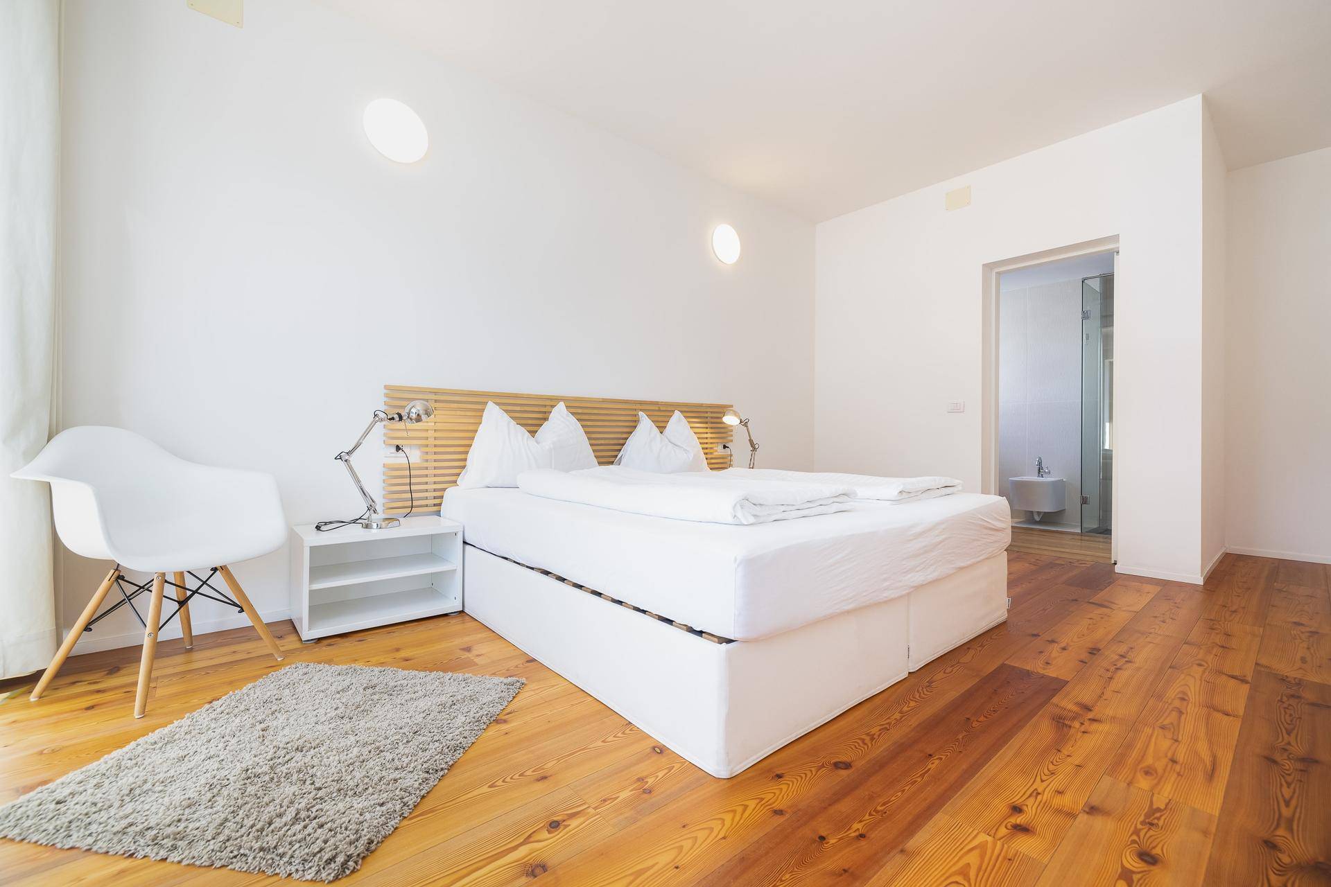 Helles Schlafzimmer mit großem weißen Bett, Holzboden und Zugang zu einem modernen Badezimmer.
