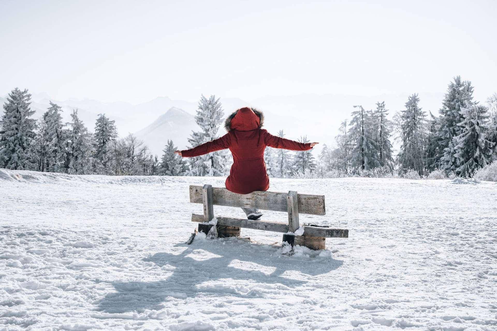 Person sitzt auf einer Holzbank in einer verschneiten Winterlandschaft mit Blick auf bewaldete Berge.