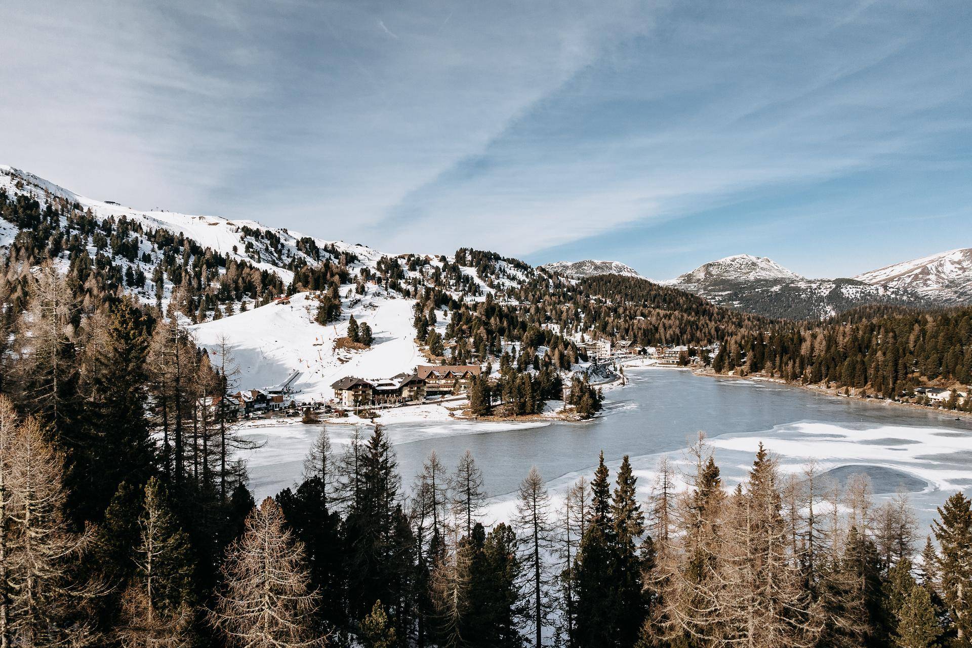 Panoramablick auf einen teilweise zugefrorenen See, umgeben von verschneiten Bergen und dichten Wäldern.