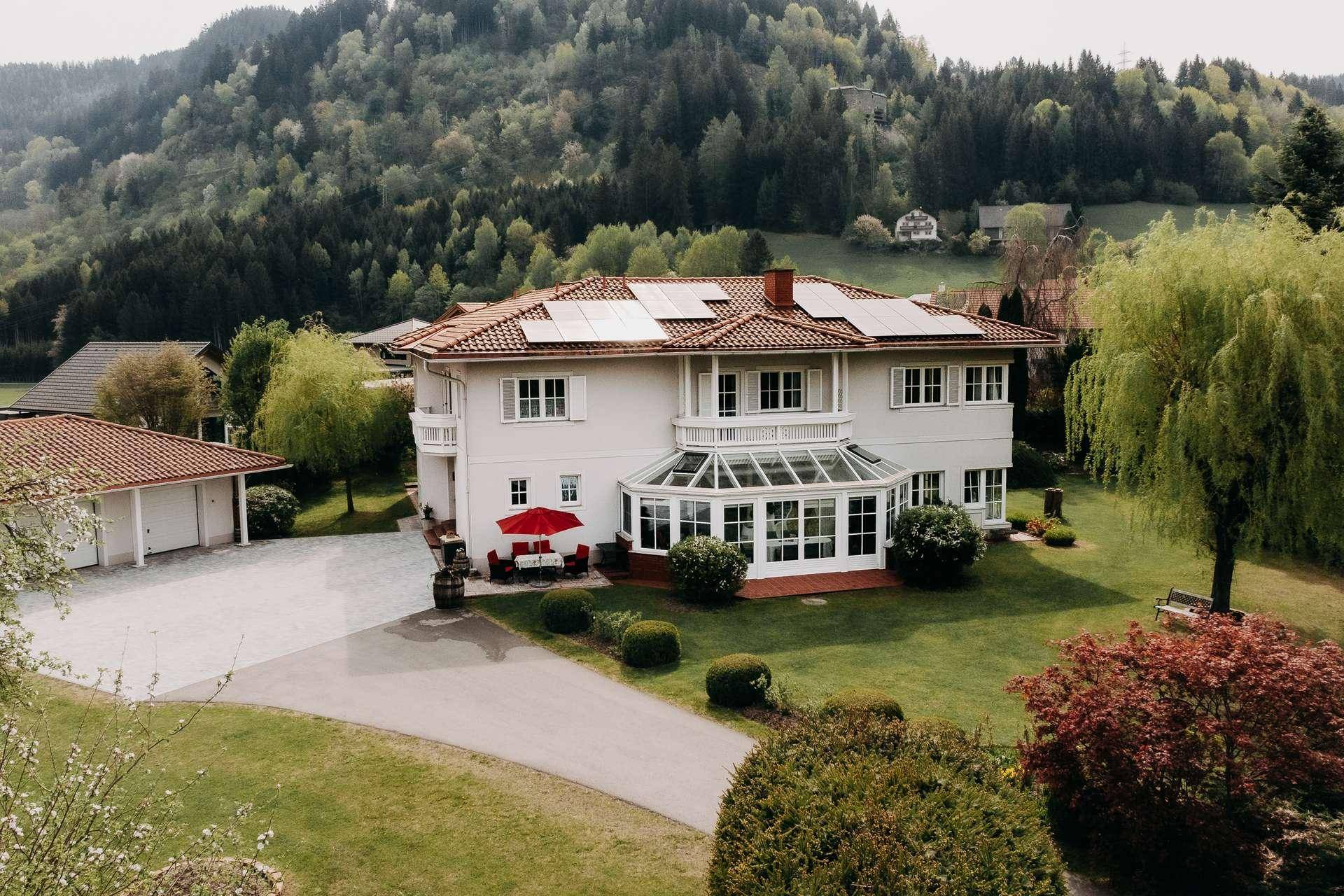 Ansicht des Hauses mit Wintergarten, Balkonen und gepflegtem Vorgarten vor bewaldetem Hügel.