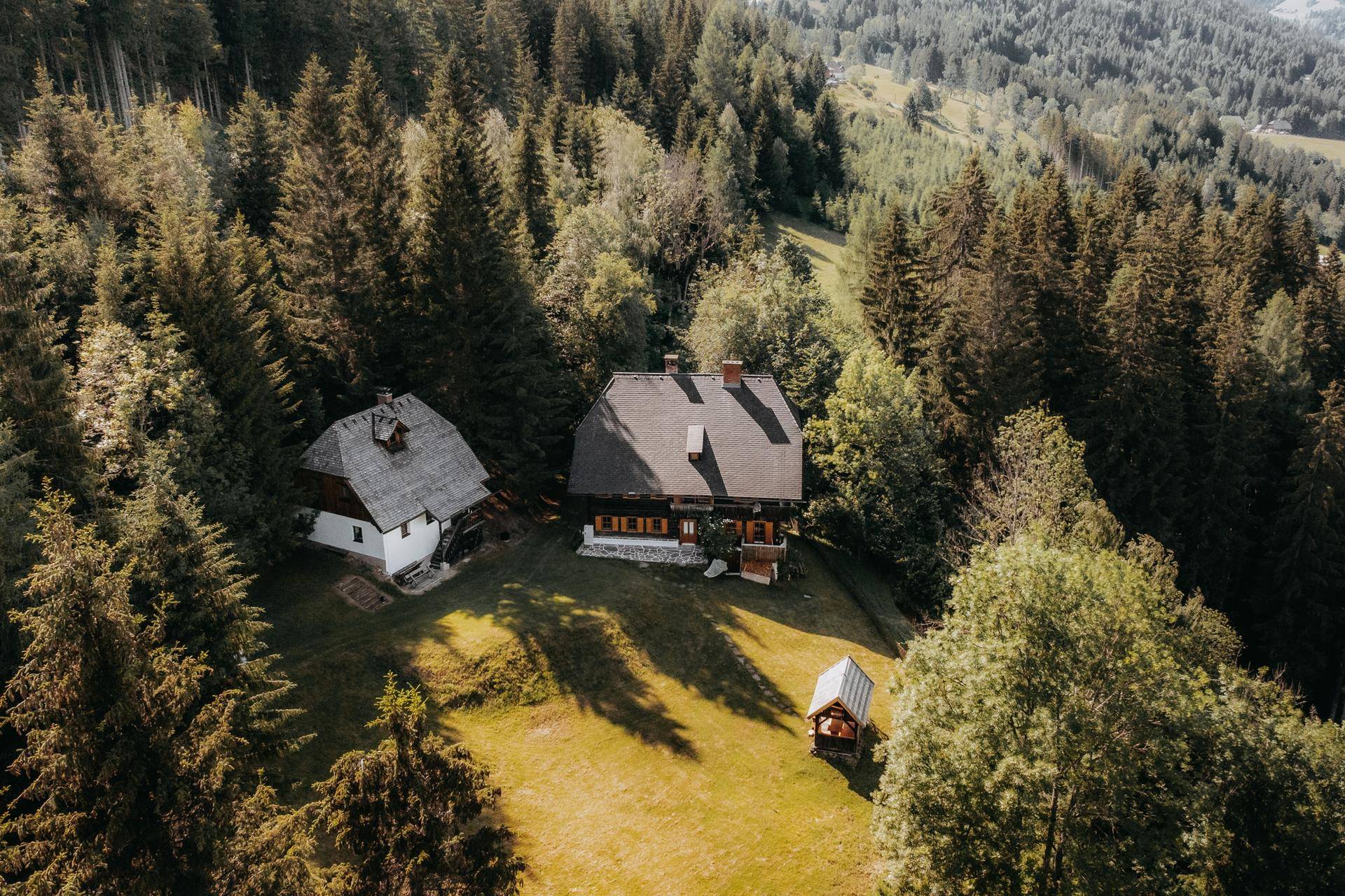 Weitläufige Luftaufnahme zweier traditioneller Häuser mit viel Grün und Wald im Hintergrund.