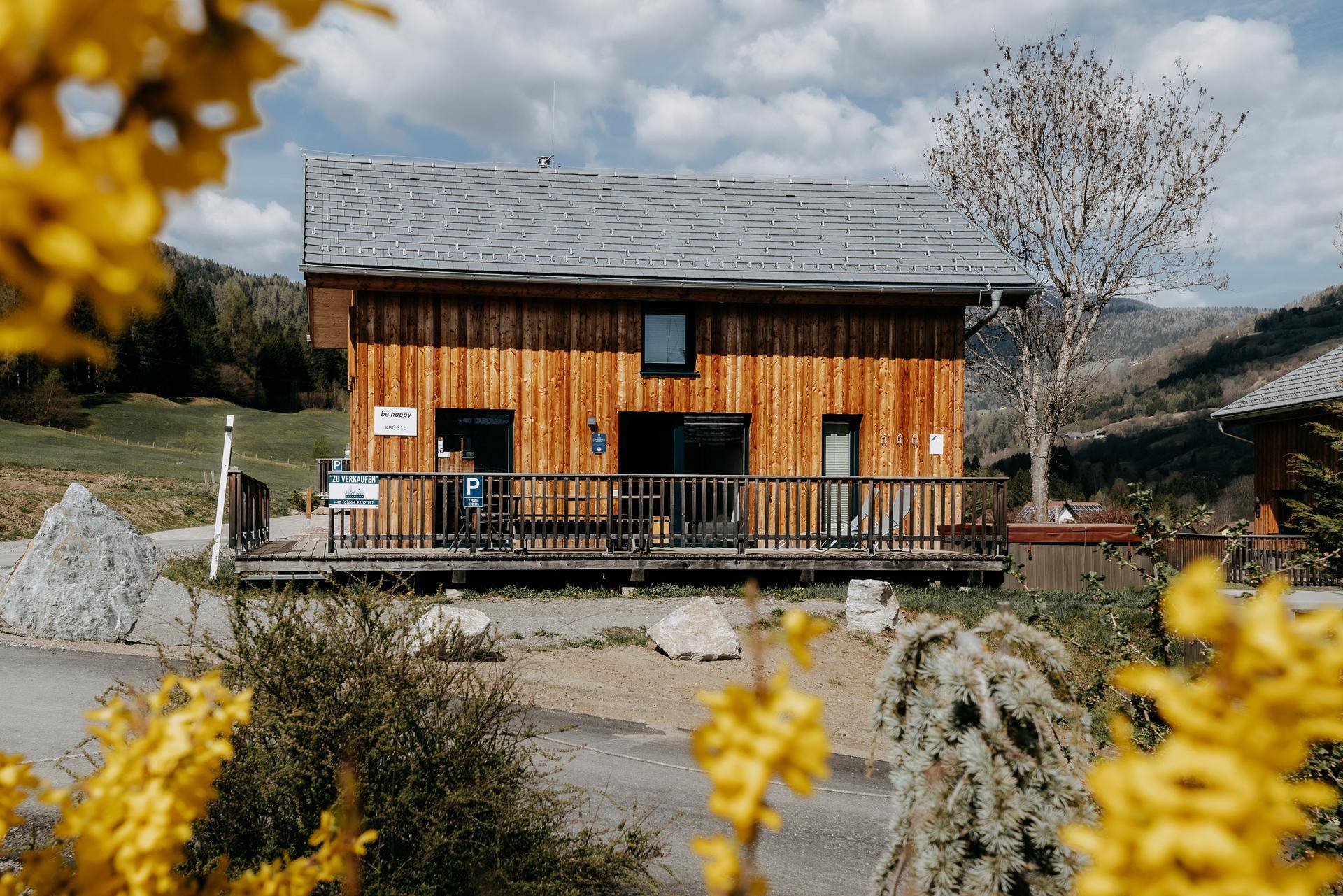 Vorderansicht eines Holzhauses mit Terrasse und Parkplatz, umgeben von blühenden Sträuchern.