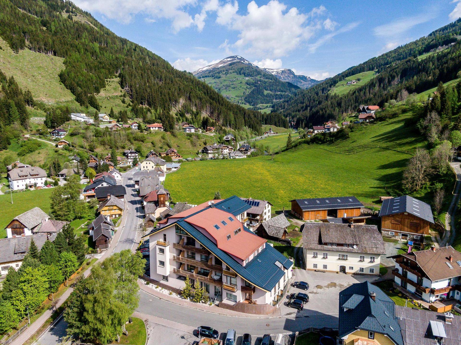 Luftaufnahme eines Dorfes in einem grünen Tal mit Bergen und einem modernen Gebäude mit Balkonen.