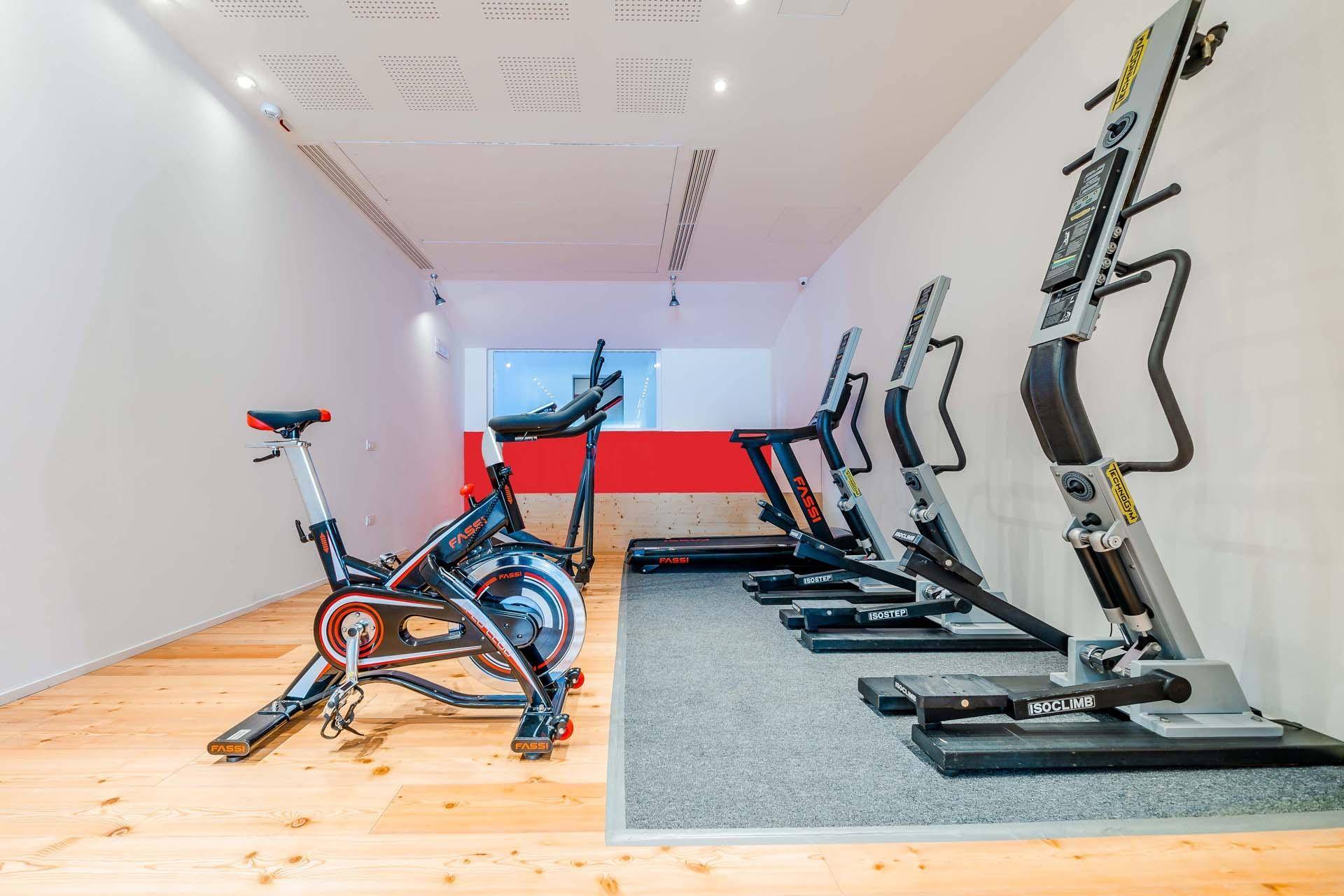 Fitnessraum mit modernen Trainingsgeräten, darunter ein Fahrrad und Klettergeräte, mit Holz- und Teppichboden.
