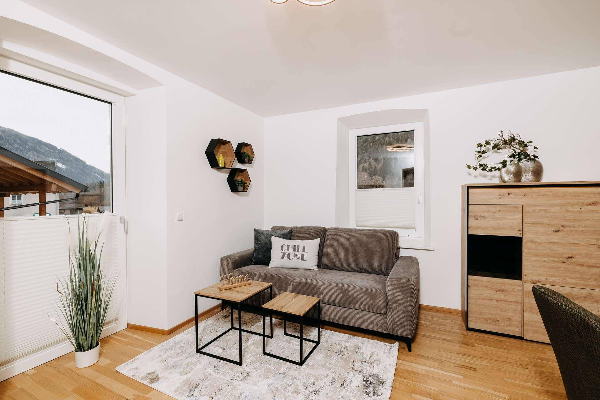 Gemütliches Wohnzimmer mit grauem Sofa, Holzboden und Blick auf die umliegende Natur.