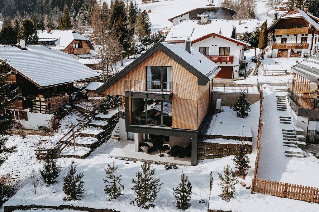 Winterliche Luftaufnahme eines modernen Holzhauses mit schneebedecktem Dach in alpiner Umgebung.