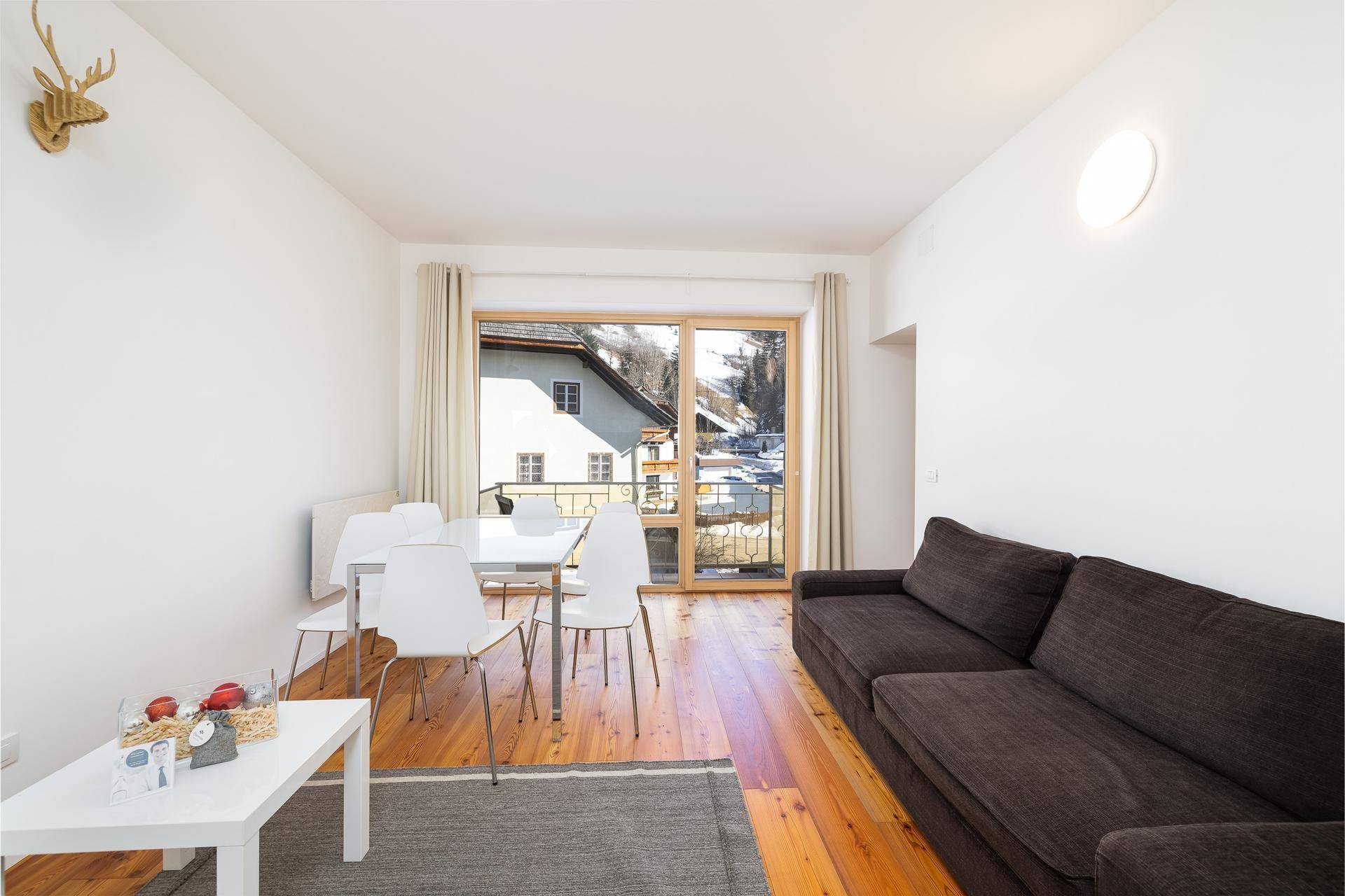 Heller Wohn-Essbereich mit dunklem Sofa, weißem Esstisch, Holzboden und Zugang zu einem Balkon.
