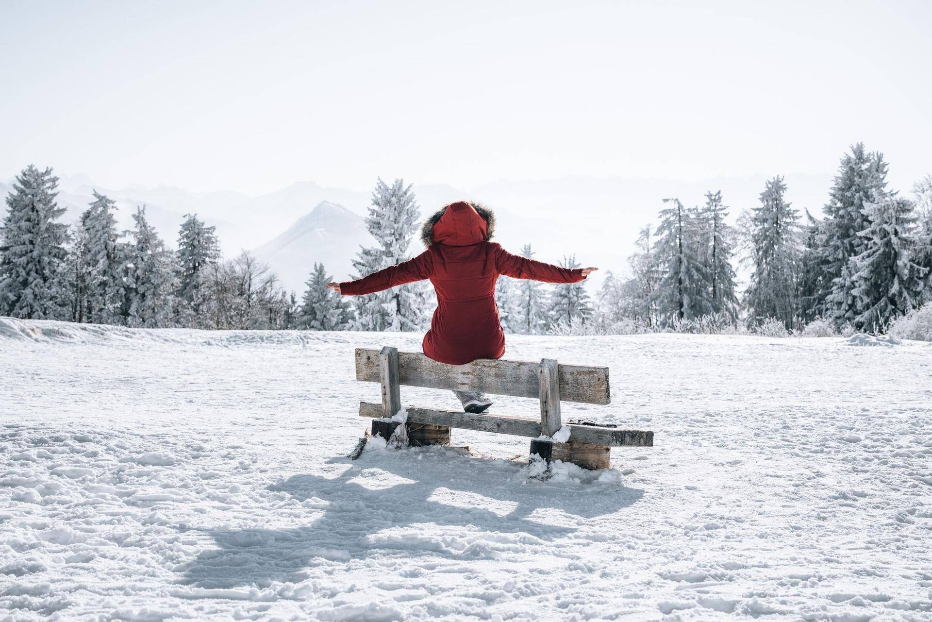 Person in roter Jacke sitzt auf einer Holzbank in einer verschneiten Winterlandschaft mit Bergen im Hintergrund.