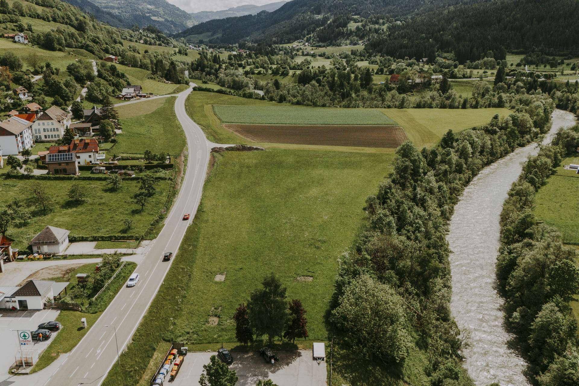 Luftaufnahme einer ländlichen Landschaft mit einem Fluss, einer Straße und weiten grünen Feldern, umgeben von Bergen.