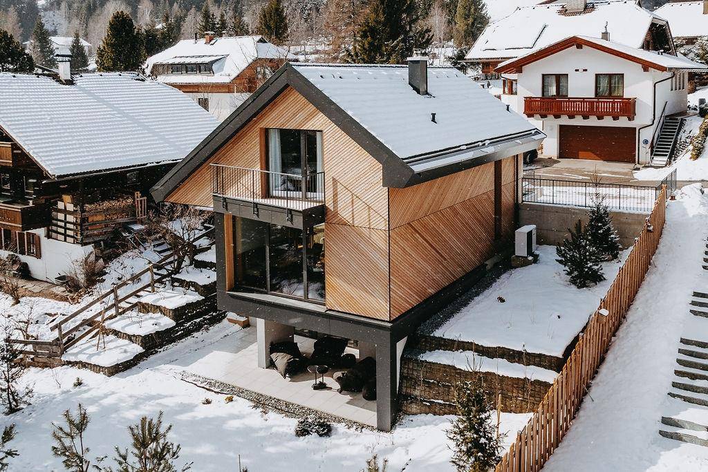 Ansicht des modernen Holzhauses im Winter mit schneebedeckter Landschaft und umliegenden Gebäuden.