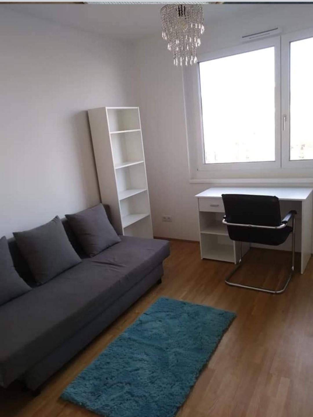 Helles Zimmer mit Schlafsofa, Schreibtisch, Regal und großem Fenster.