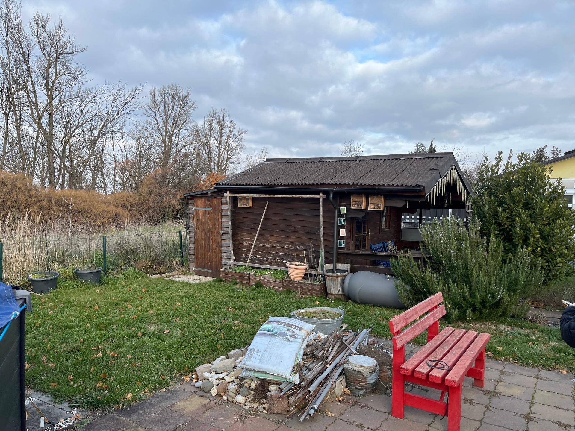 Garten mit Rasenfläche, einem Holzschuppen und einer roten Gartenbank unter bewölktem Himmel.