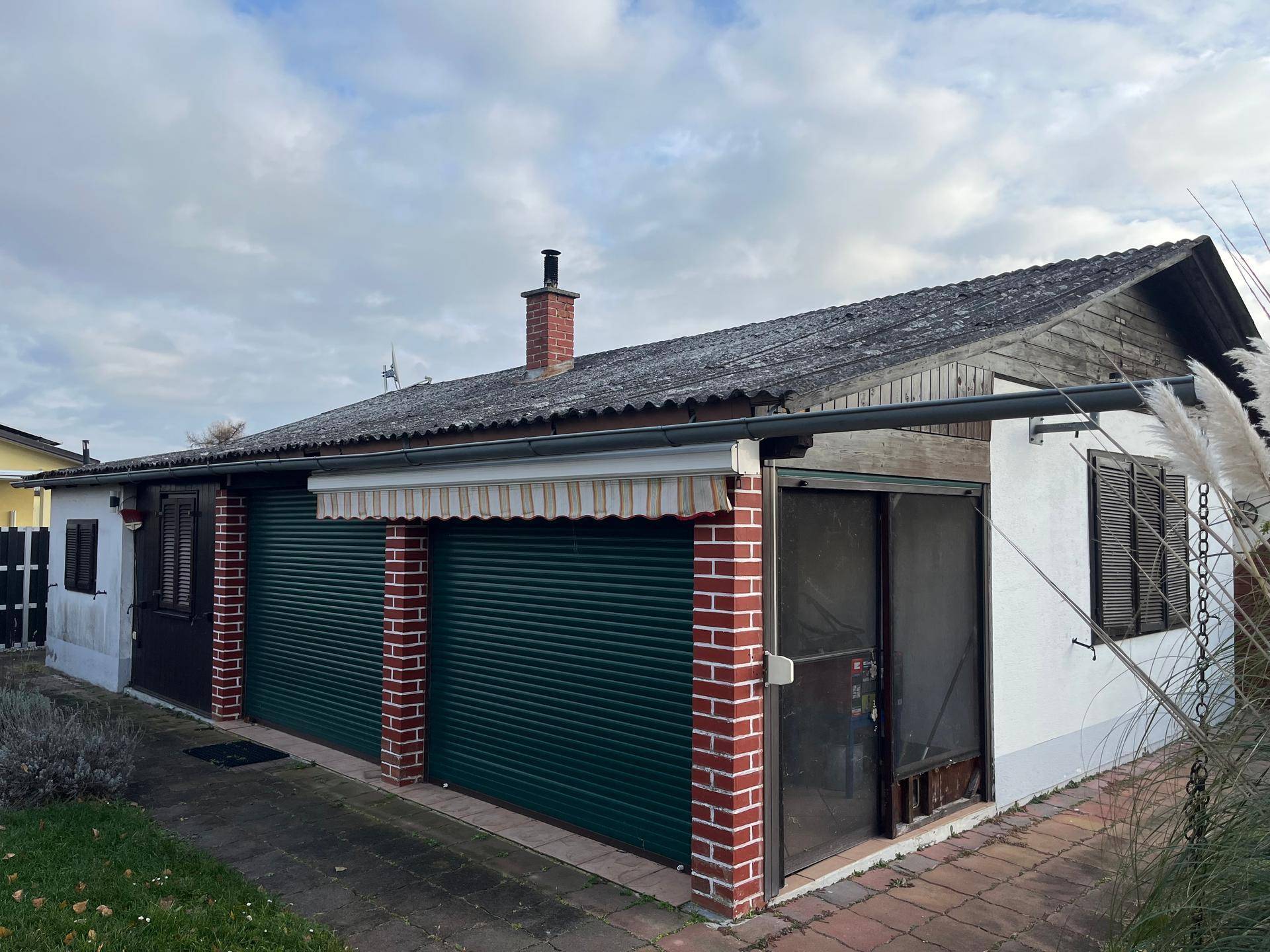 Seitenansicht des Hauses mit grünen Rollläden, Ziegelpfeilern und einem Vordach unter bewölktem Himmel.