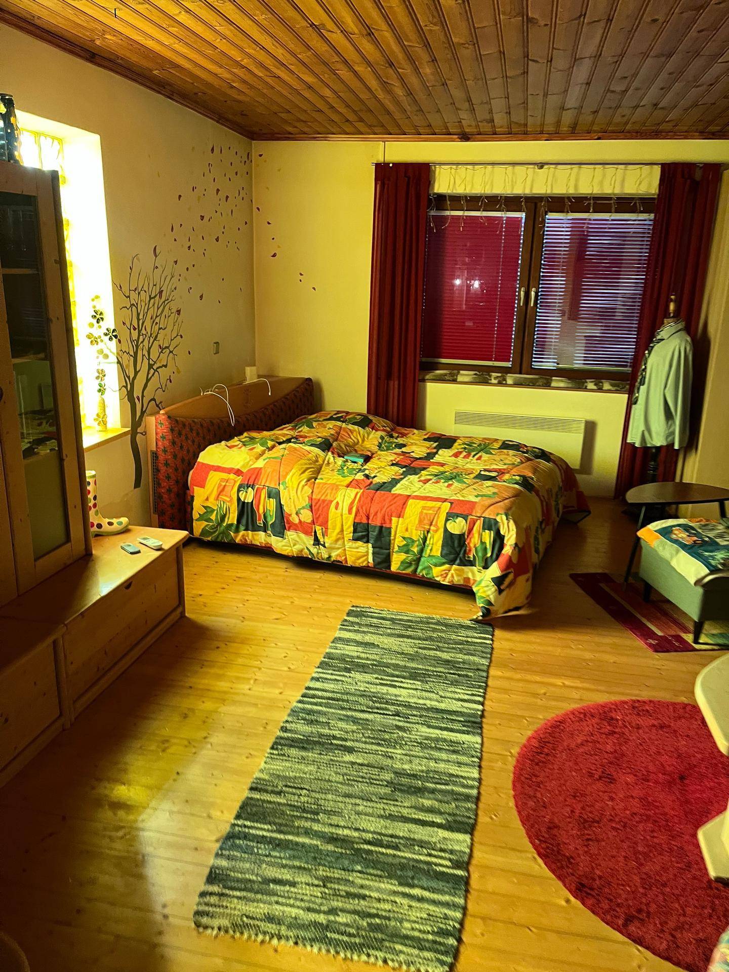 Schlafzimmer mit bunter Tagesdecke, Holzdecke und Fenster mit roten Vorhängen und Jalousien.