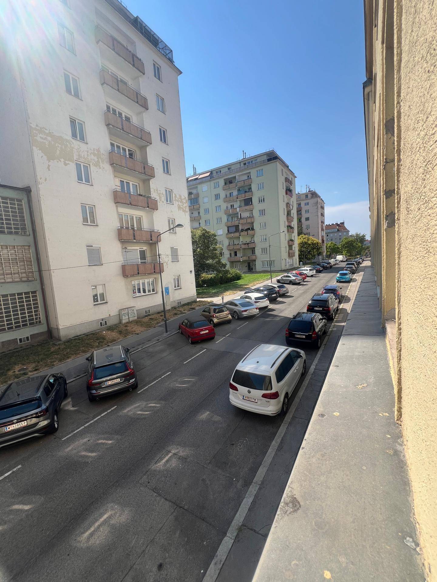 Weitläufiger Blick auf eine städtische Straße mit Gebäuden und parkenden Fahrzeugen an einem sonnigen Tag.