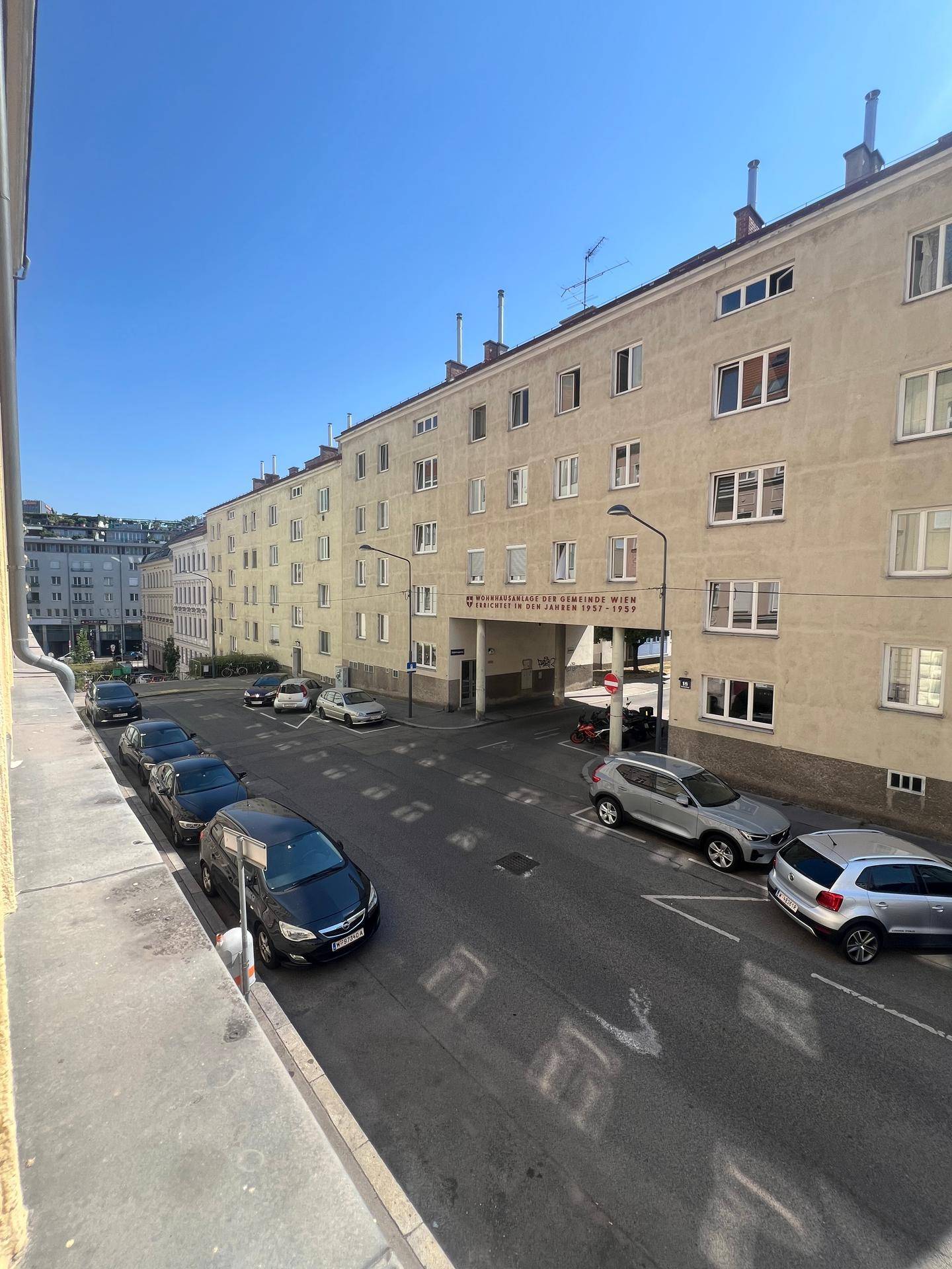 Blick auf eine belebte Straße mit parkenden Autos und einem mehrstöckigen Wohngebäude unter blauem Himmel.