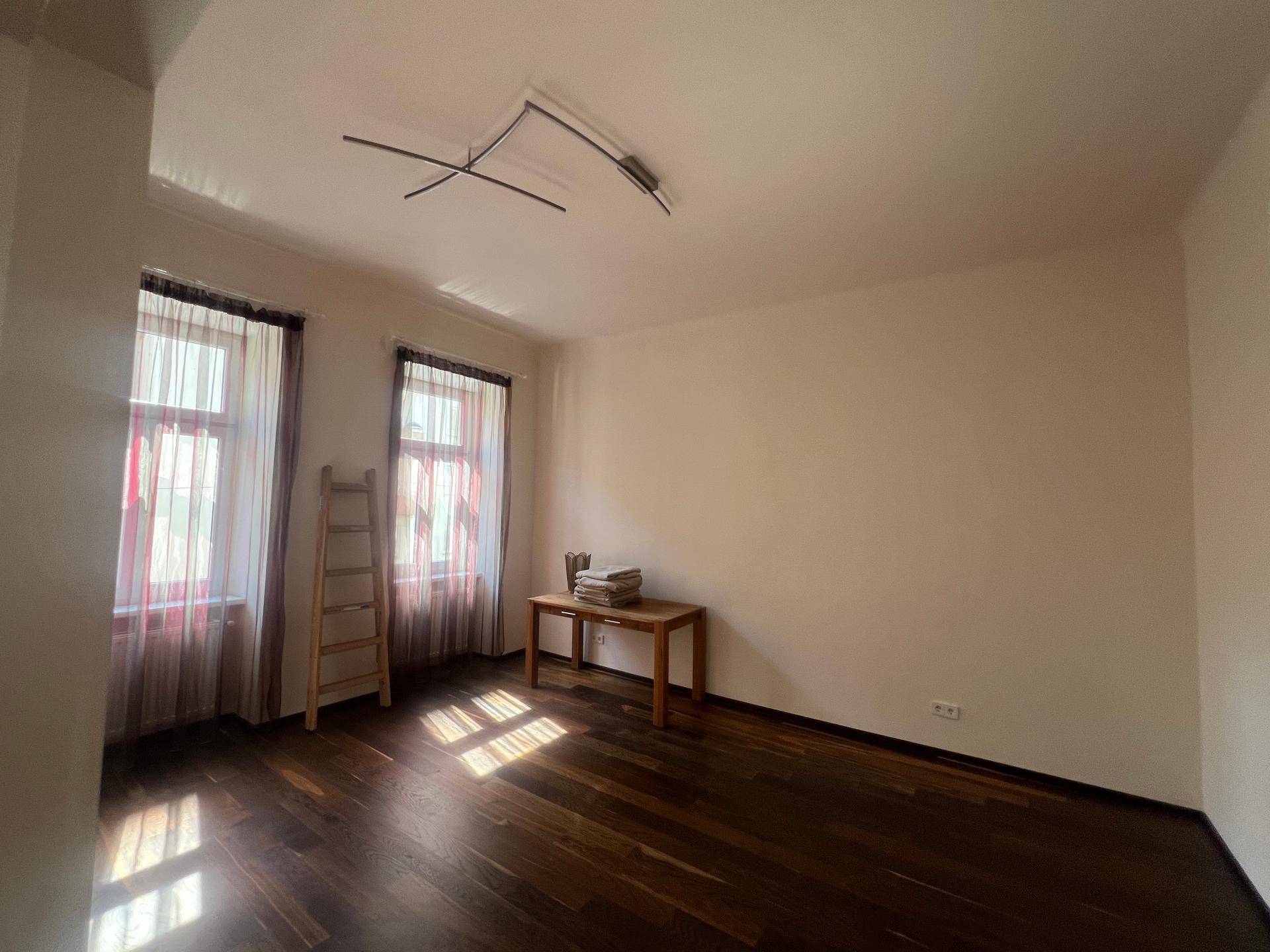Heller Raum mit dunklem Holzboden, einem kleinen Tisch und natürlichem Lichteinfall durch die Fenster.