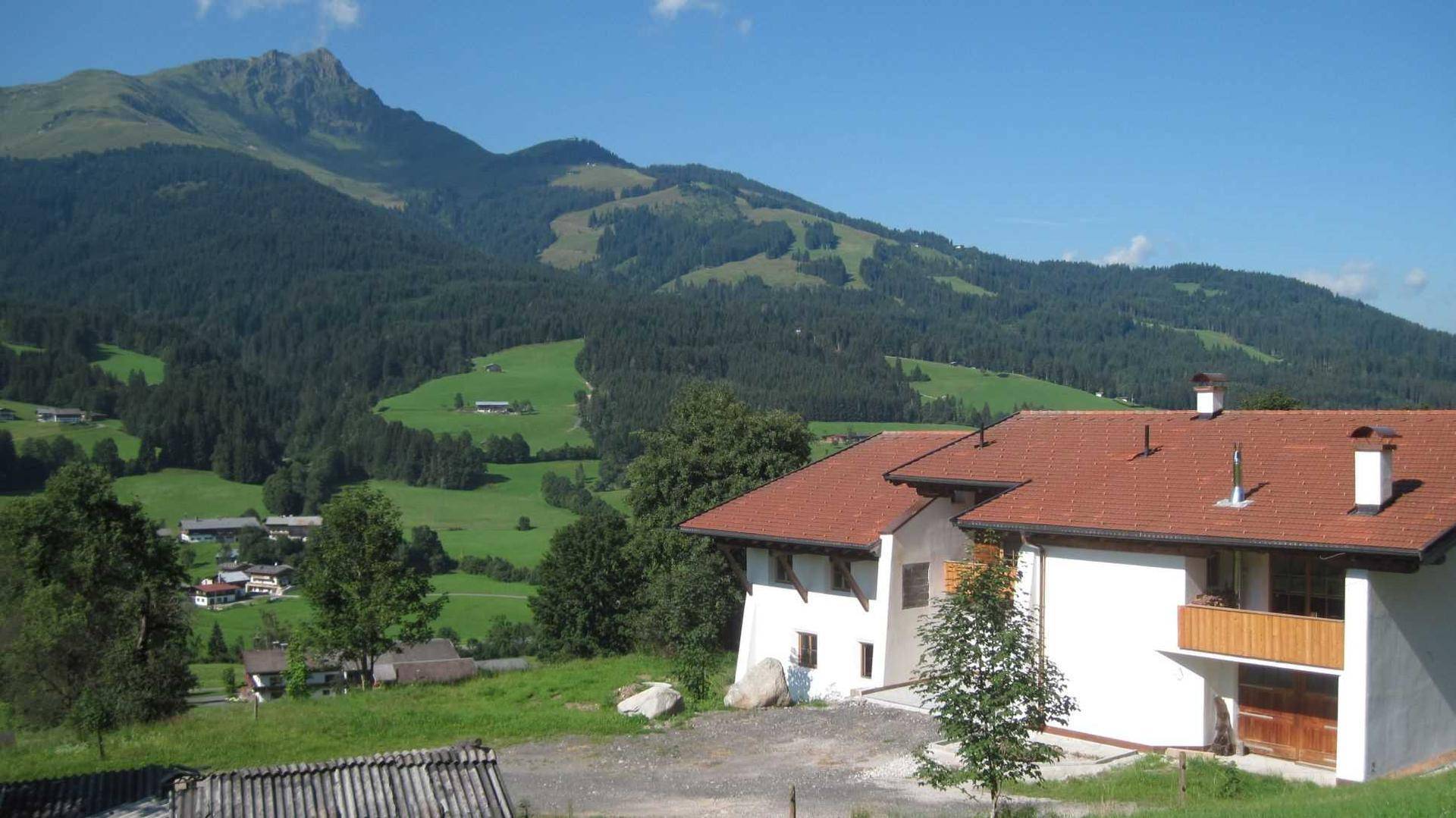 Weißes Haus mit rotem Ziegeldach und Holzbalkon vor einer Kulisse aus grünen Hügeln und Bergen.