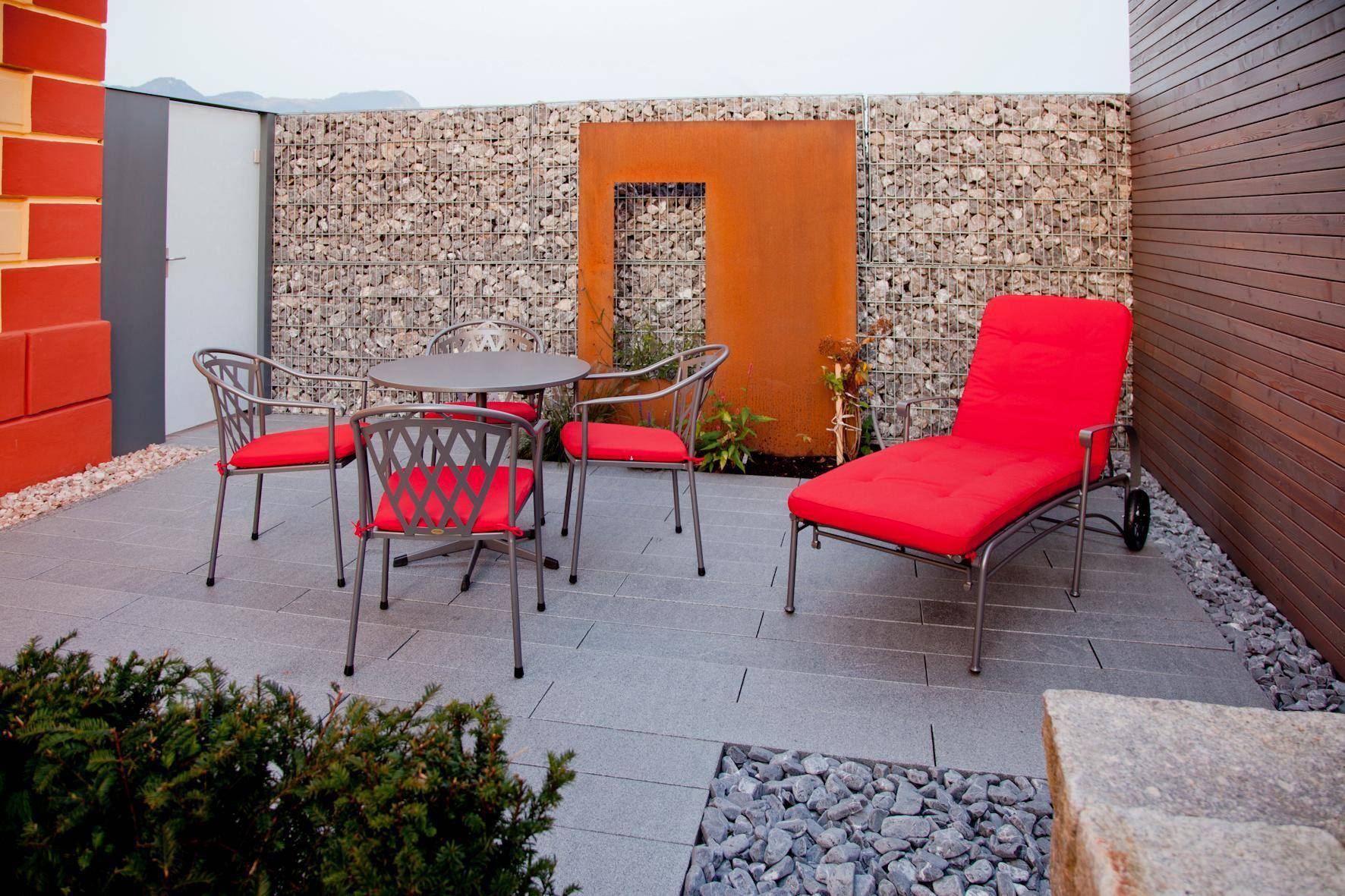 Moderne Terrasse mit Sitzgelegenheiten, Liegestuhl und Gabionenwand im Außenbereich.