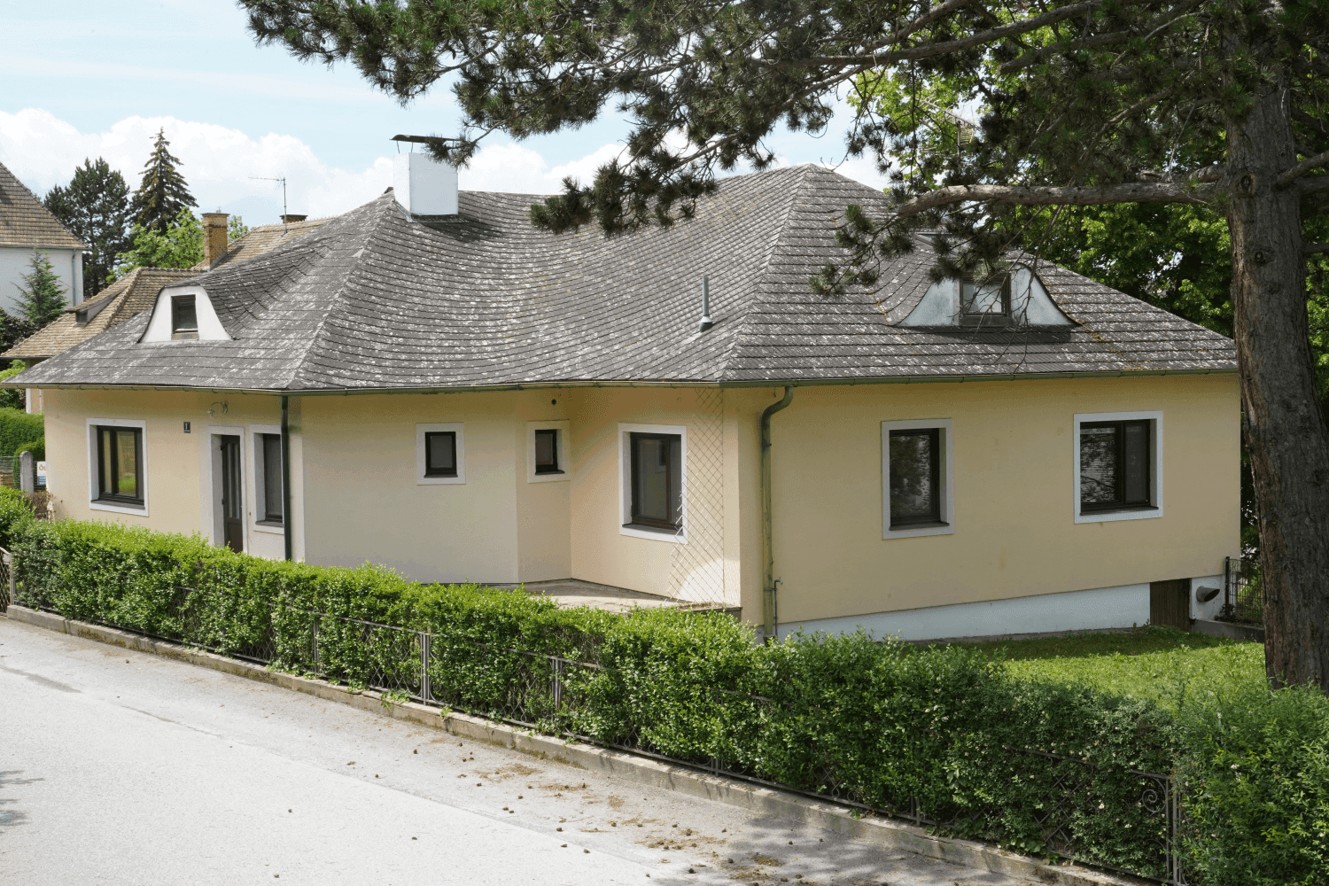 Seitenansicht des Hauses mit gepflegter Hecke und schattenspendenden Bäumen im Gartenbereich.
