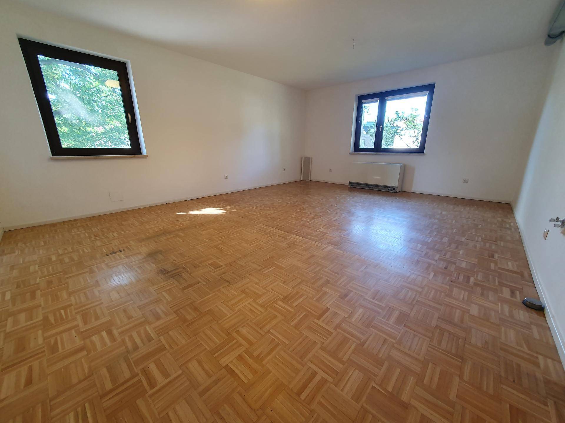Geräumiges, leeres Zimmer mit Parkettboden und zwei großen Fenstern für viel Tageslicht.