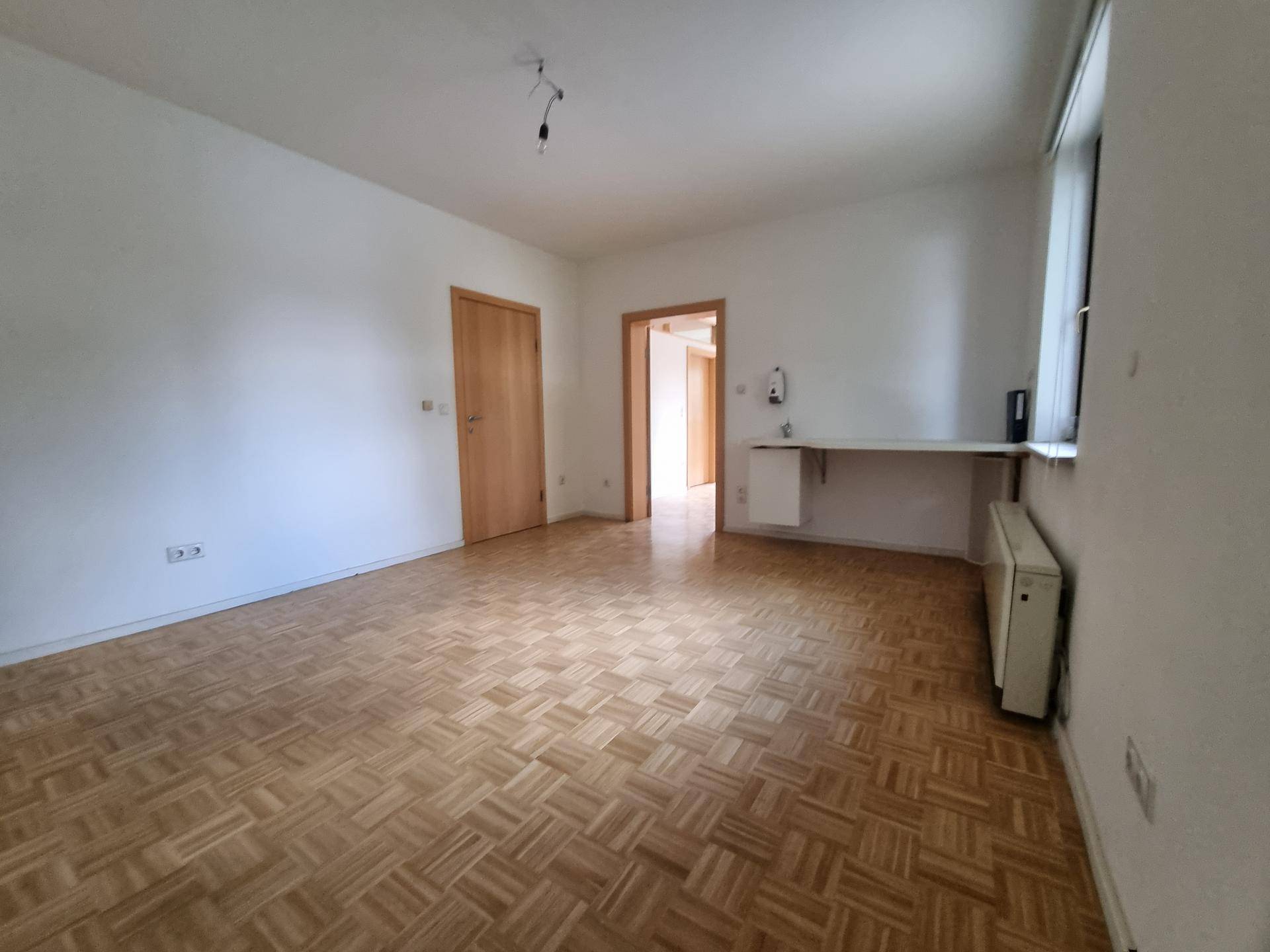 Heller Raum mit Parkettboden und einer kleinen Ablagefläche unter dem Fenster.