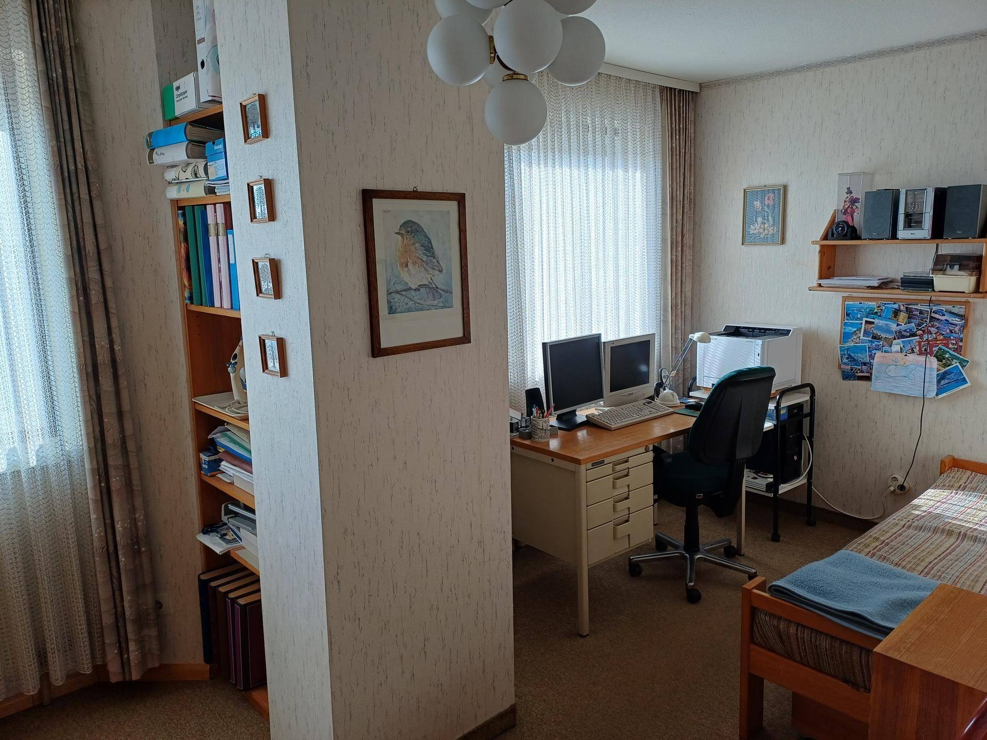 Arbeitszimmer mit Schreibtisch, Computern, Bücherregal und einem Einzelbett.