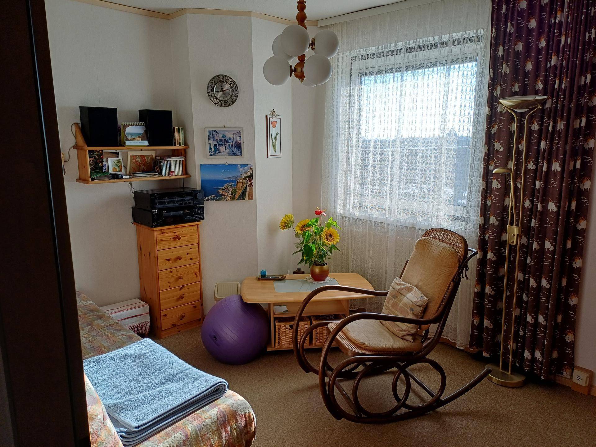 Schlafzimmer mit Einzelbett, Schaukelstuhl, kleinem Tisch und Fenster mit Vorhängen.