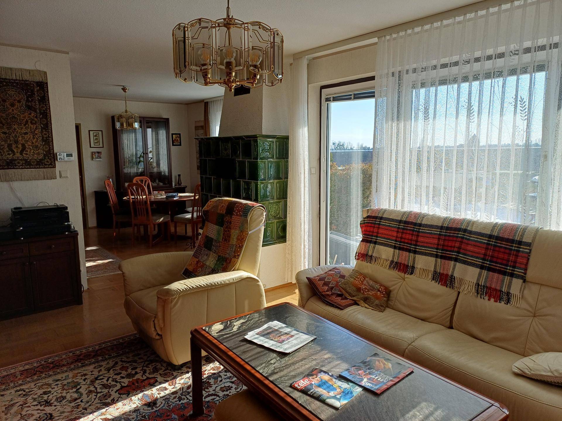 Helles Wohnzimmer mit beigem Sofa, traditionellem Kachelofen und großem Fenster mit Vorhängen.