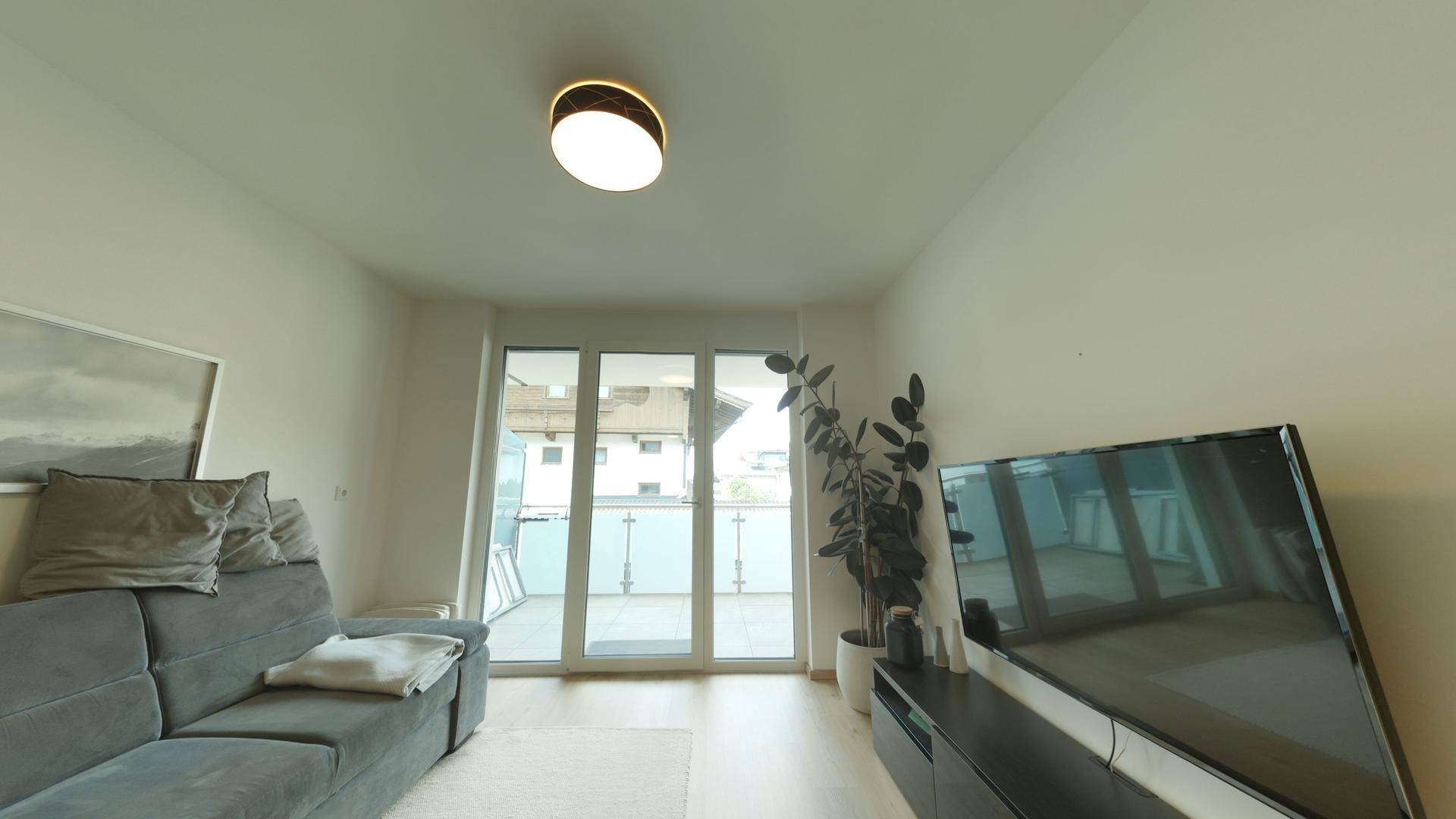 Gemütliches Wohnzimmer mit grauem Sofa, Fernseher und Zugang zu einem Balkon.