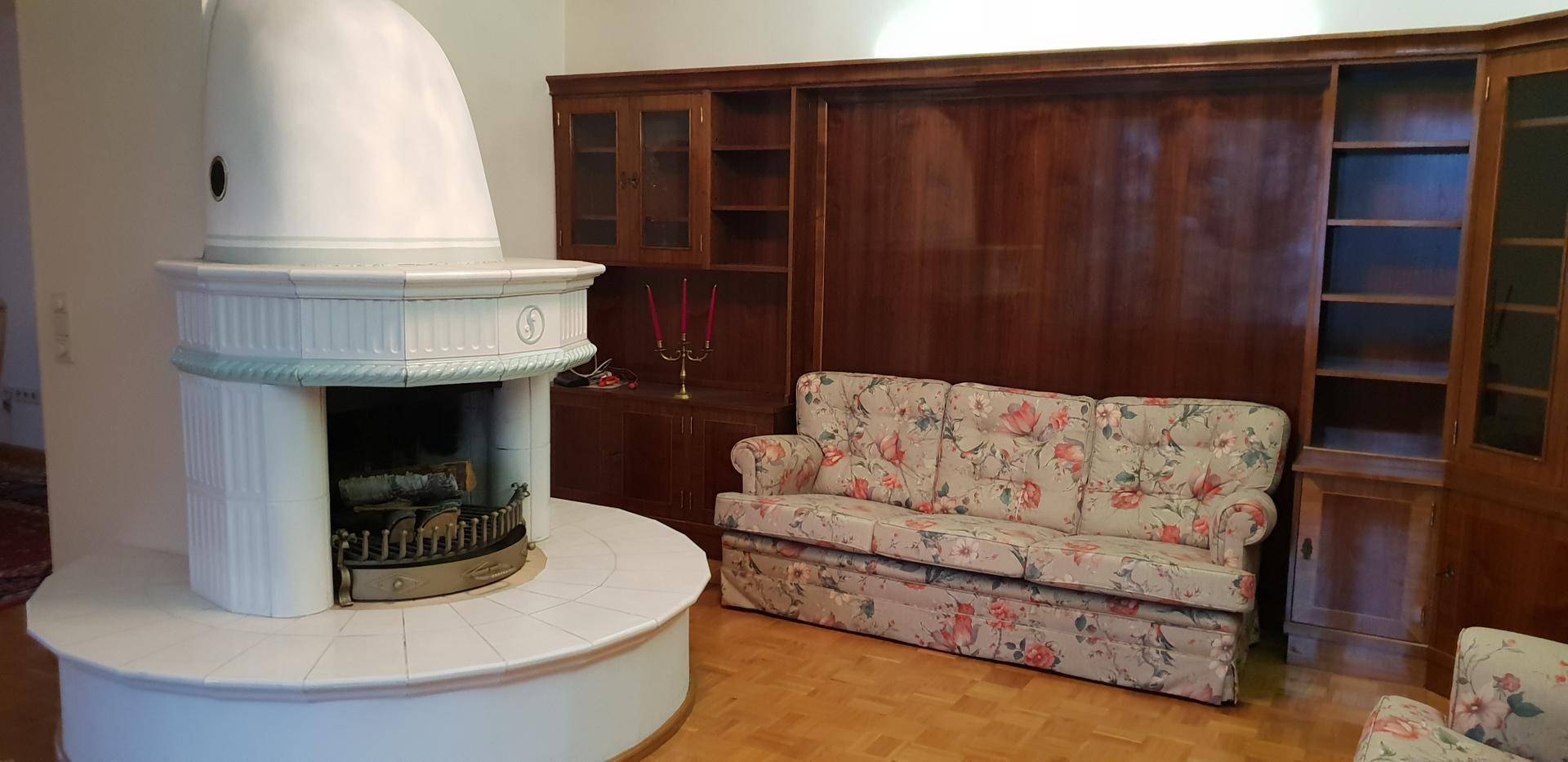 Gemütliches Wohnzimmer mit traditionellem Kachelofen, floralem Sofa und Holzeinbauten.