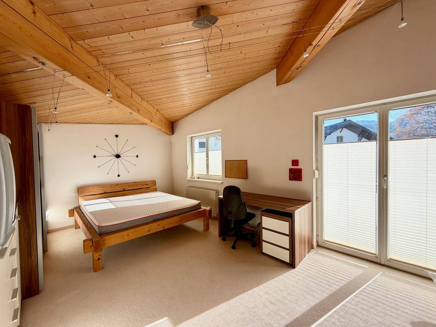 Geräumiges Schlafzimmer mit Holzbalkendecke, Teppichboden und Zugang zum Balkon.