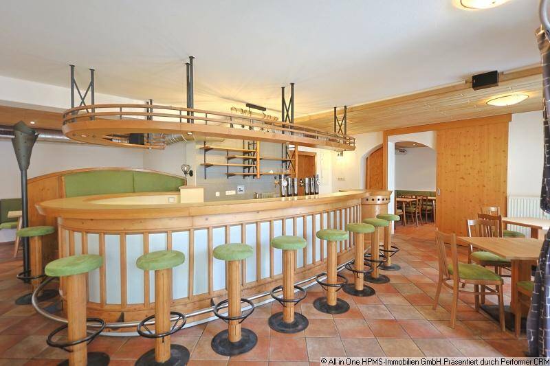 Thekenbereich mit Holzverkleidung, Barhockern und einem Hängeregal über der Theke, ergänzt durch Holzbalken an der Decke.