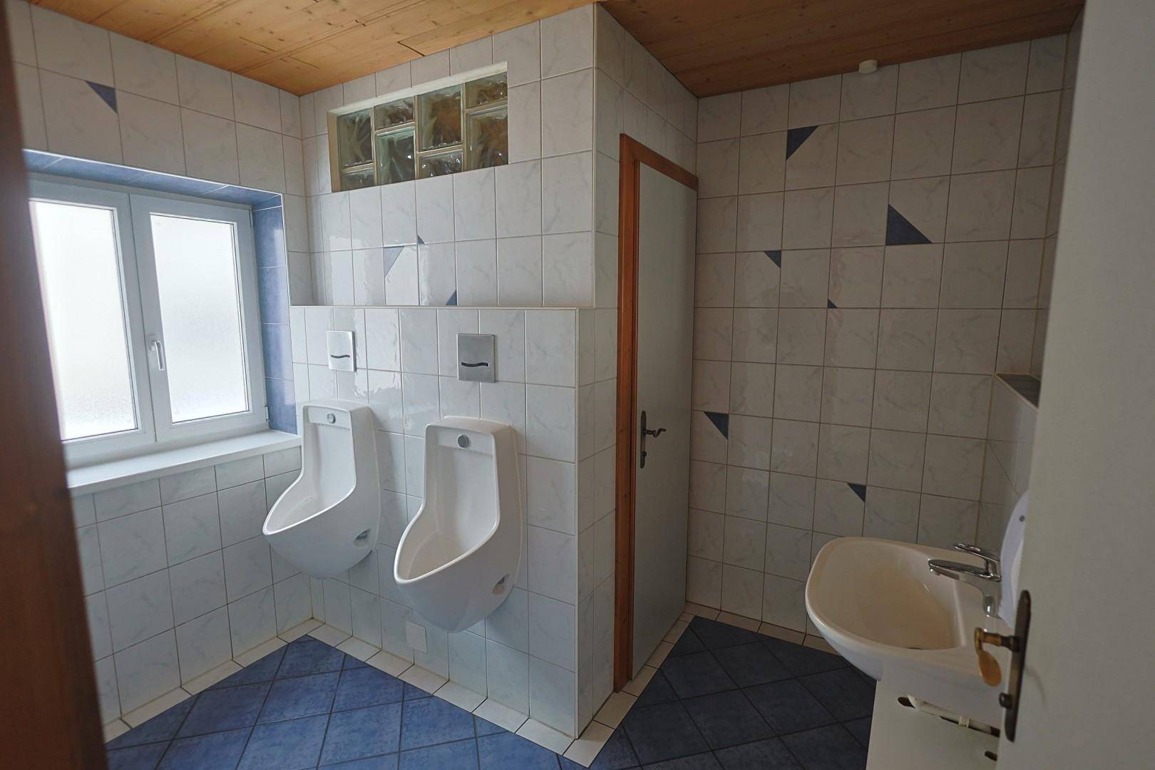 Herrenwaschraum mit zwei Urinalen, Waschbecken, weißen Fliesen und einem Fenster für natürliche Belüftung.