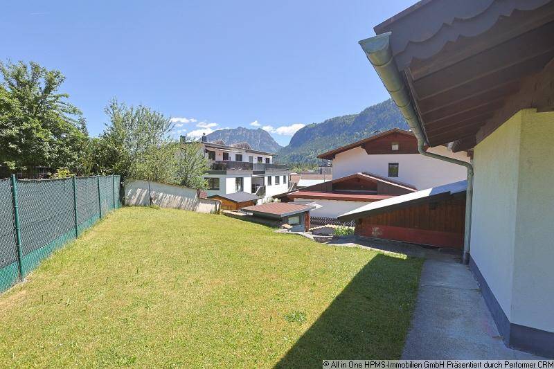 Rückseite des Hauses mit Rasenfläche, einem Weg und Blick auf weitere Gebäude und Berge.