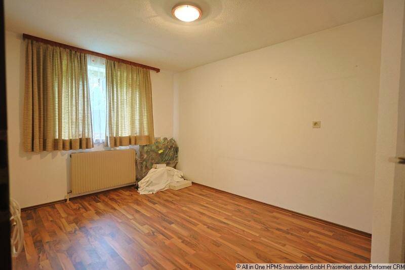 Einfaches Schlafzimmer mit Holzboden, weißen Wänden und einem Fenster mit Vorhängen.