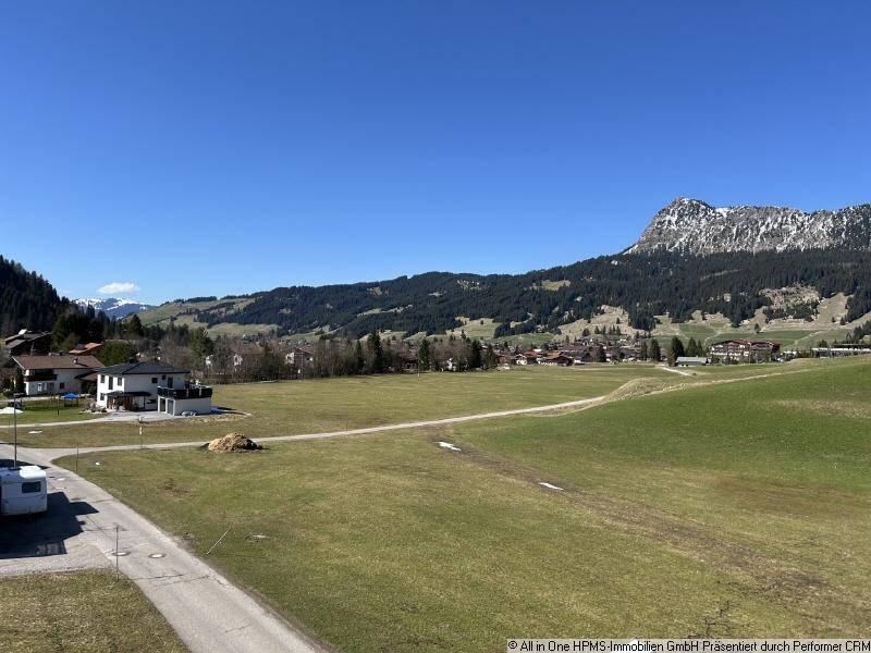 Weitläufige grüne Landschaft mit Bergen im Hintergrund und einigen Häusern in der Ferne.