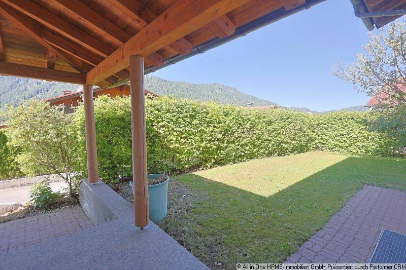 Überdachter Außenbereich mit Holzbalken, Blick auf eine kleine Rasenfläche und eine dichte Hecke.