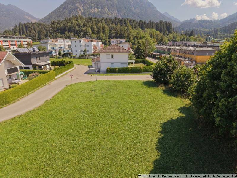 Luftaufnahme eines grünen Baugrundstücks mit Blick auf die umliegenden Berge und Wohnhäuser.