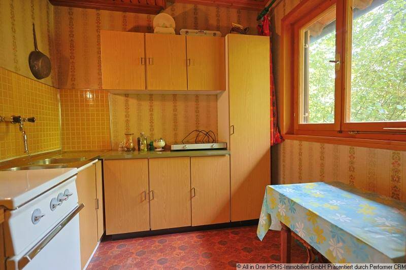 Einfache Küche mit gelben Fliesen, älteren Holzschränken und einem Fenster mit Waldblick.