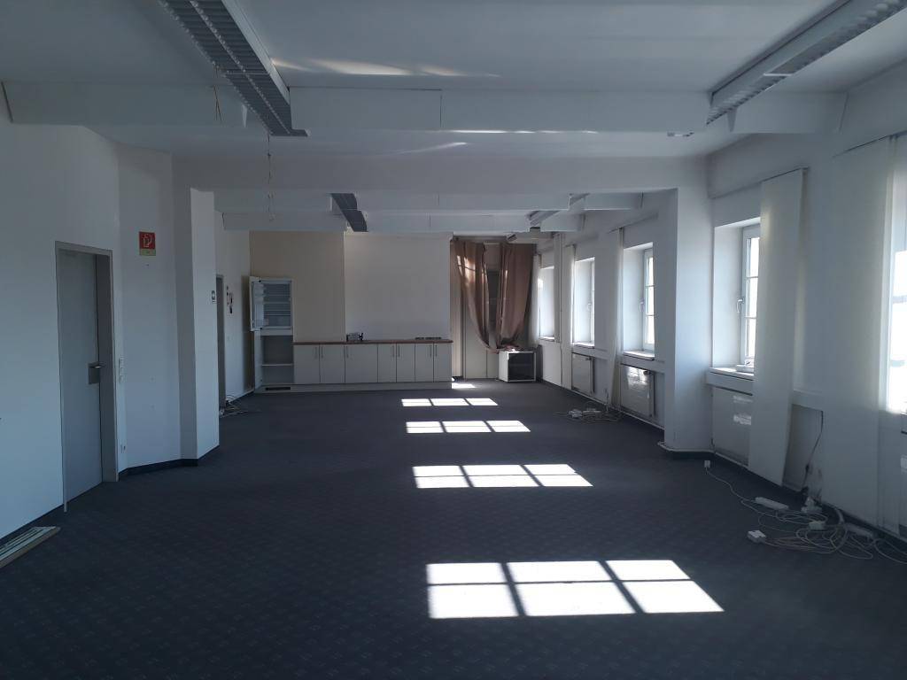 Großer, heller Büroraum mit Teppichboden, hohen Decken und viel natürlichem Licht durch die Fenster.