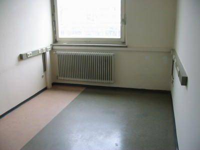 Ecke eines Raumes mit Fenster, Heizkörper und zwei verschiedenen Bodenbelägen, die Renovierungsbedarf andeuten.